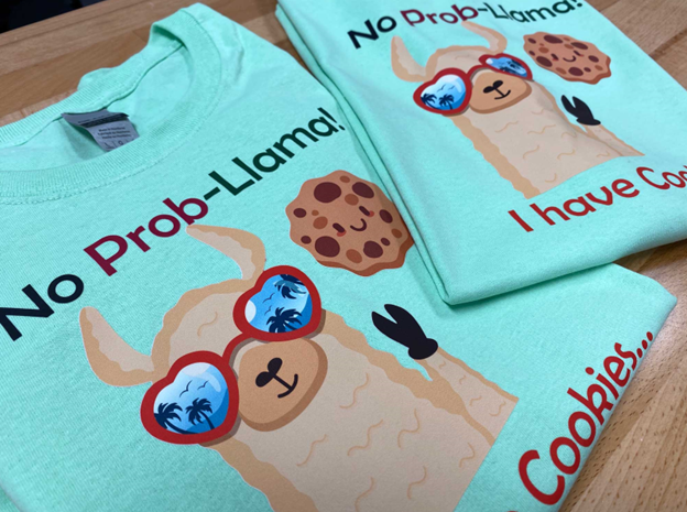 Image of a teal t-shirt that features a cartoon llama wearing heart sunglasses with a cookie and the text "no prob-llama!"